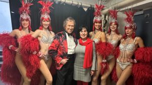 Bezirksbürgermeisterin Emine Demirbüken-Wegner und Entertainer Bert Beel im rot-schwarzen Sakko zwischen fünf Can-Can-Tänzerinnen mit roten Federkostümen und Kopfschmuck.