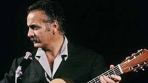 Historisches Farbfoto: Ein Mann mit grauem Haar und Schnurrbart in dunklem Sakko spielt vor schwarzem Hintergrund Gitarre an einem Mikrofon und blickt nach links.