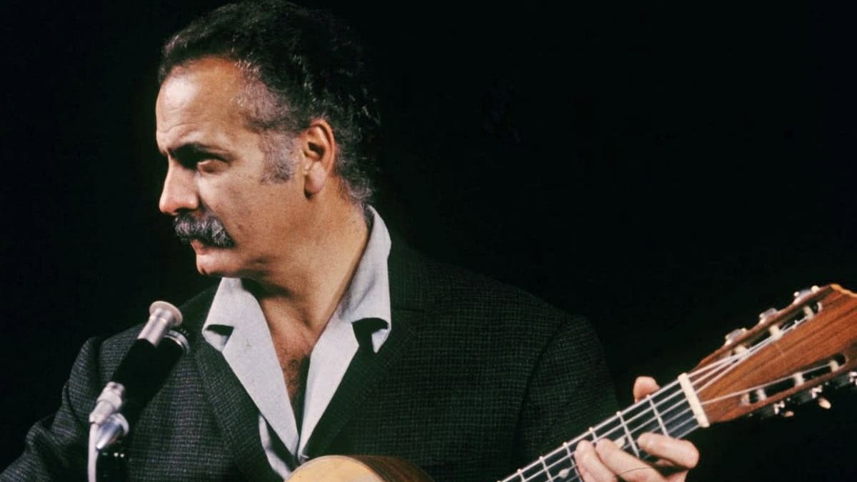 Historisches Farbfoto: Ein Mann mit grauem Haar und Schnurrbart in dunklem Sakko spielt vor schwarzem Hintergrund Gitarre an einem Mikrofon und blickt nach links.