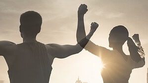 Grafik in Sepiatönen: Ein Mann und eine Frau stehen mit dem Rücken zum Betrachter vor einer Stadtsilhouette im Gegenlicht und recken die Fäuste in die Höhe, unten rechts der Schriftzug „Team Gewaltmanagement, Coach für Zivilcourage“.