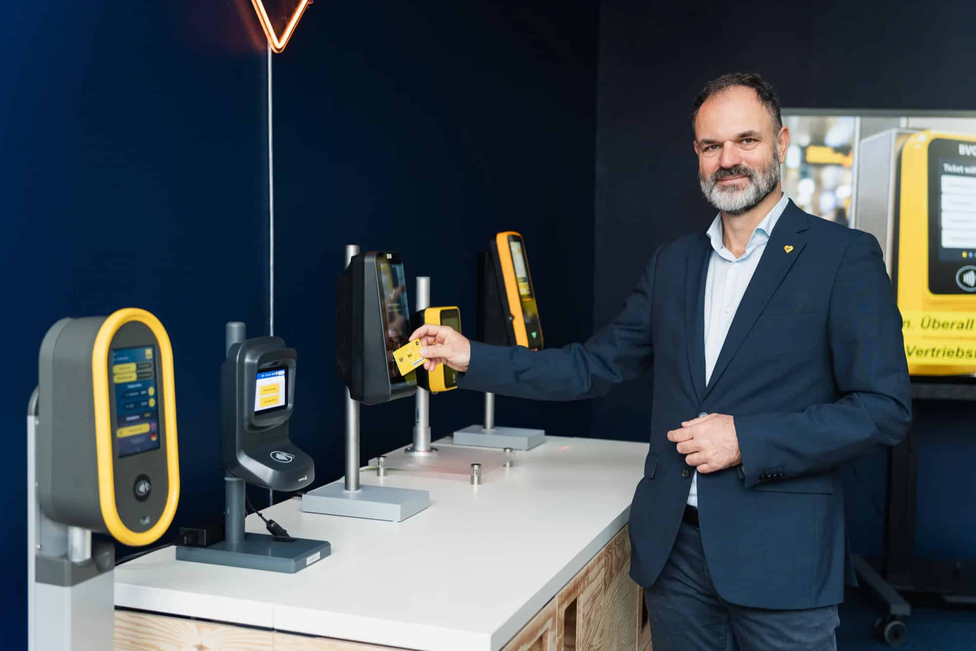 Ein Mann mit grauem Bart im dunklen Sakko hält eine gelbe Karte an einen von mehreren Validatoren, die auf einem weißen Tisch vor einer dunkelblauen Wand stehen