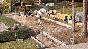 Baustelle mit Bauzaun am Waldrand, ein Arbeiter steht auf der abgezogenen Fläche, daneben ein gelber Minibagger und Holzpaletten.