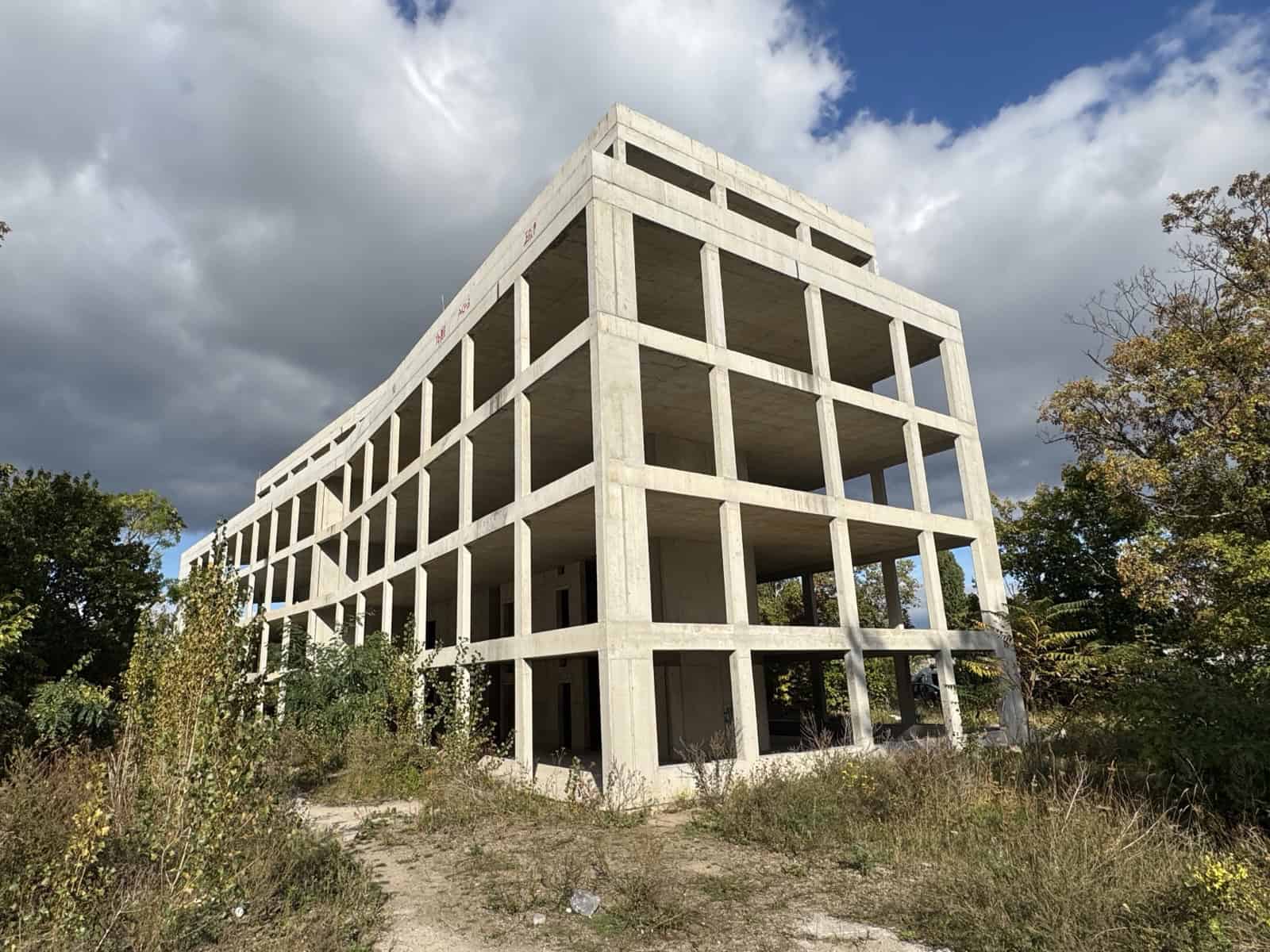 Unverputzter Betonrohbau eines mehrgeschossigen Gebäudes ohne Fenster und Fassade, umgeben von Bäumen, Sträuchern und hohem Gras, davor ein Trampelpfad