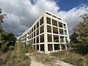 Unverputzter Betonrohbau eines mehrgeschossigen Gebäudes ohne Fenster und Fassade, umgeben von Bäumen, Sträuchern und hohem Gras, davor ein Trampelpfad