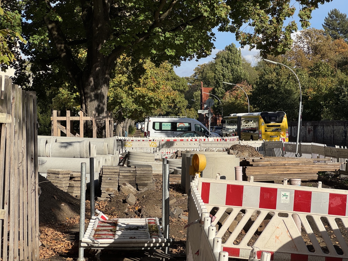 Baustelle mit gelagerten Betonrohren, Schachtringen, Pflastersteinen und Holzbohlen hinter rot-weißen Absperrgittern, im Hintergrund ein gelber BVG-Bus