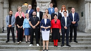 Gruppenfoto von 15 Frauen und Männern in Anzug, Kostüm oder legerer Kleidung, die in drei Reihen auf einer Steintreppe vor einem verzierten Gebäudeportal mit zwei Rundbögen stehen