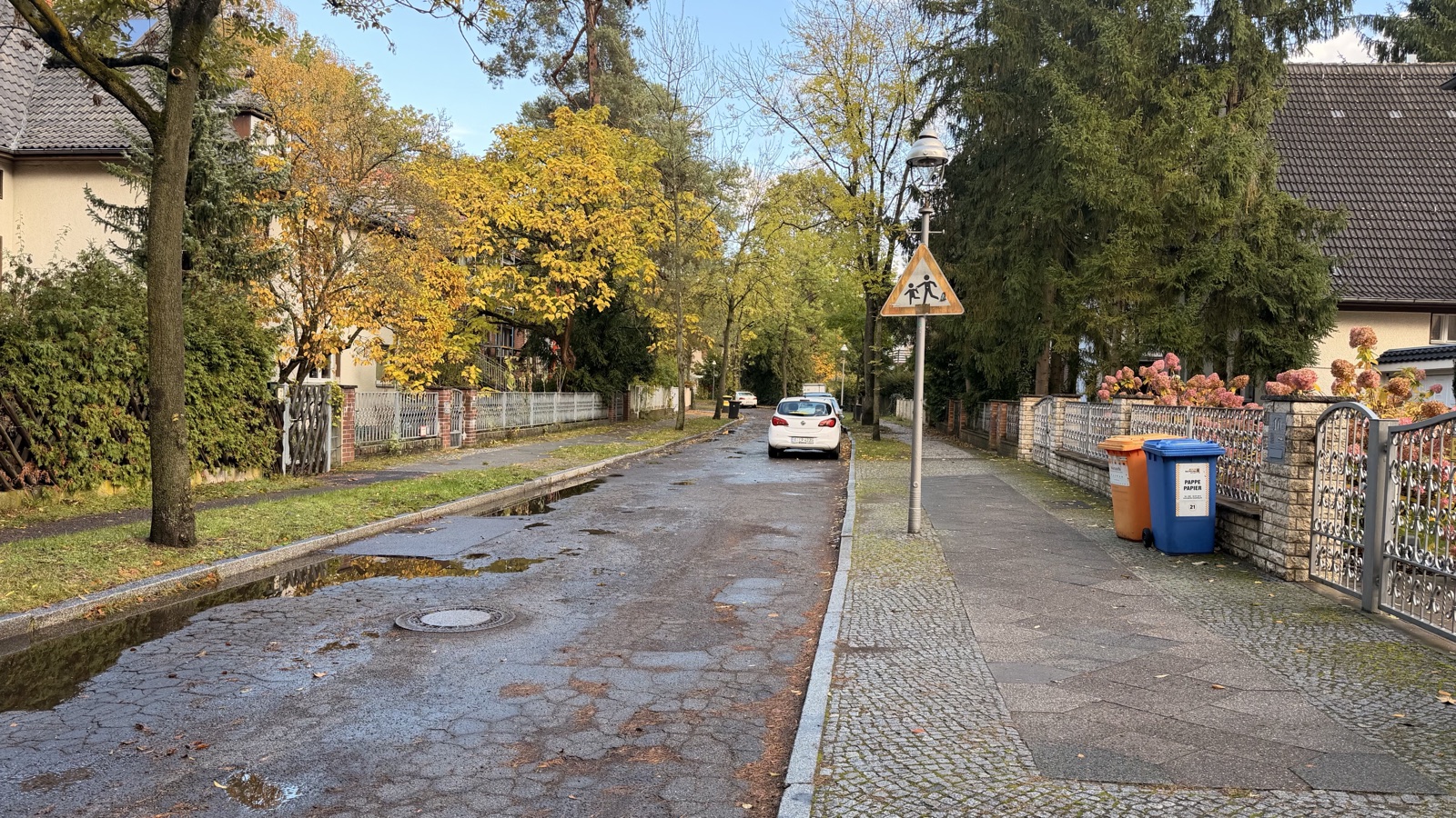 Fuchssteinerweg in Frohnau im Herbst: rissige, geflickte Fahrbahn mit Pfützen, Gehweg mit Laterne und Schild Achtung Kinder, Einfamilienhäuser und Bäume mit Herbstlaub