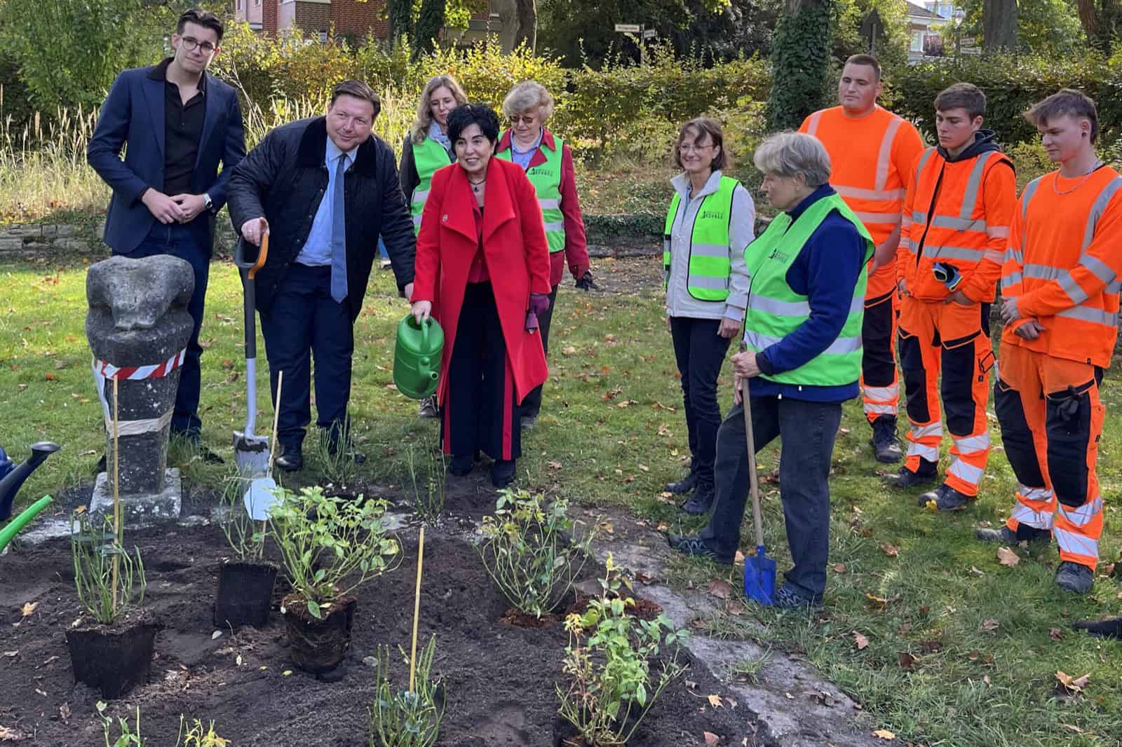 Eine Gruppe von Erwachsenen, teils in grünen Warnwesten, teils in orangefarbener Arbeitskleidung, steht auf einer Rasenfläche unter herbstlichen Bäumen an einem frisch angelegten Beet mit jungen Pflanzen; links eine steinerne Figur, eine Frau im roten Mantel hält eine grüne Gießkanne.