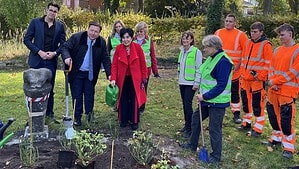 Eine Gruppe von Erwachsenen, teils in grünen Warnwesten, teils in orangefarbener Arbeitskleidung, steht auf einer Rasenfläche unter herbstlichen Bäumen an einem frisch angelegten Beet mit jungen Pflanzen; links eine steinerne Figur, eine Frau im roten Mantel hält eine grüne Gießkanne.
