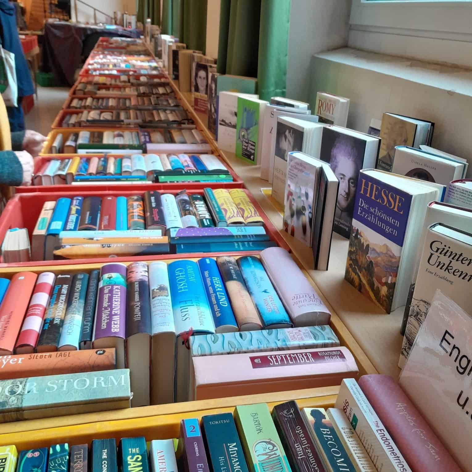 Lange Reihen roter und gelber Kisten voller Bücher auf Tischen in einem Saal, an der Fensterbank stehen weitere Bücher aufgestellt.