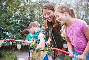 Eine Betreuerin untersucht mit mehreren Kindern am Ufer eines Gewässers mit Keschern das Leben in einem Teich.