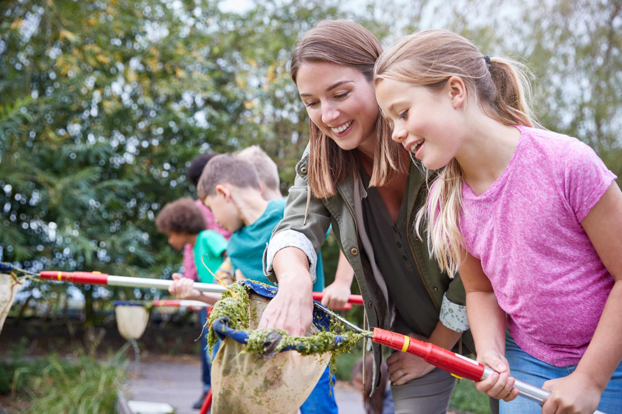 Eine Betreuerin untersucht mit mehreren Kindern am Ufer eines Gewässers mit Keschern das Leben in einem Teich.
