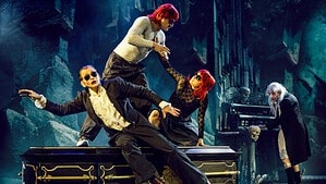 Bühnenszene: Ein Mann mit Rüschenhemd, Sonnenbrille und rot geschminkten Lippen lehnt auf einem schwarzen Sarg, dahinter zwei Figuren mit roten Perücken, rechts eine Gestalt mit langen grauen Haaren an einer Orgel mit Totenschädeln