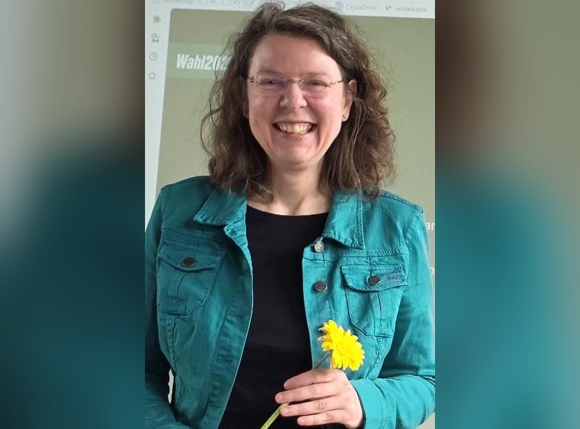 Eine Frau mit Brille und türkisem Jäckchen hält vor einer Leinwand mit Abstimmungsergebnissen eine gelbe Blume in den Händen.