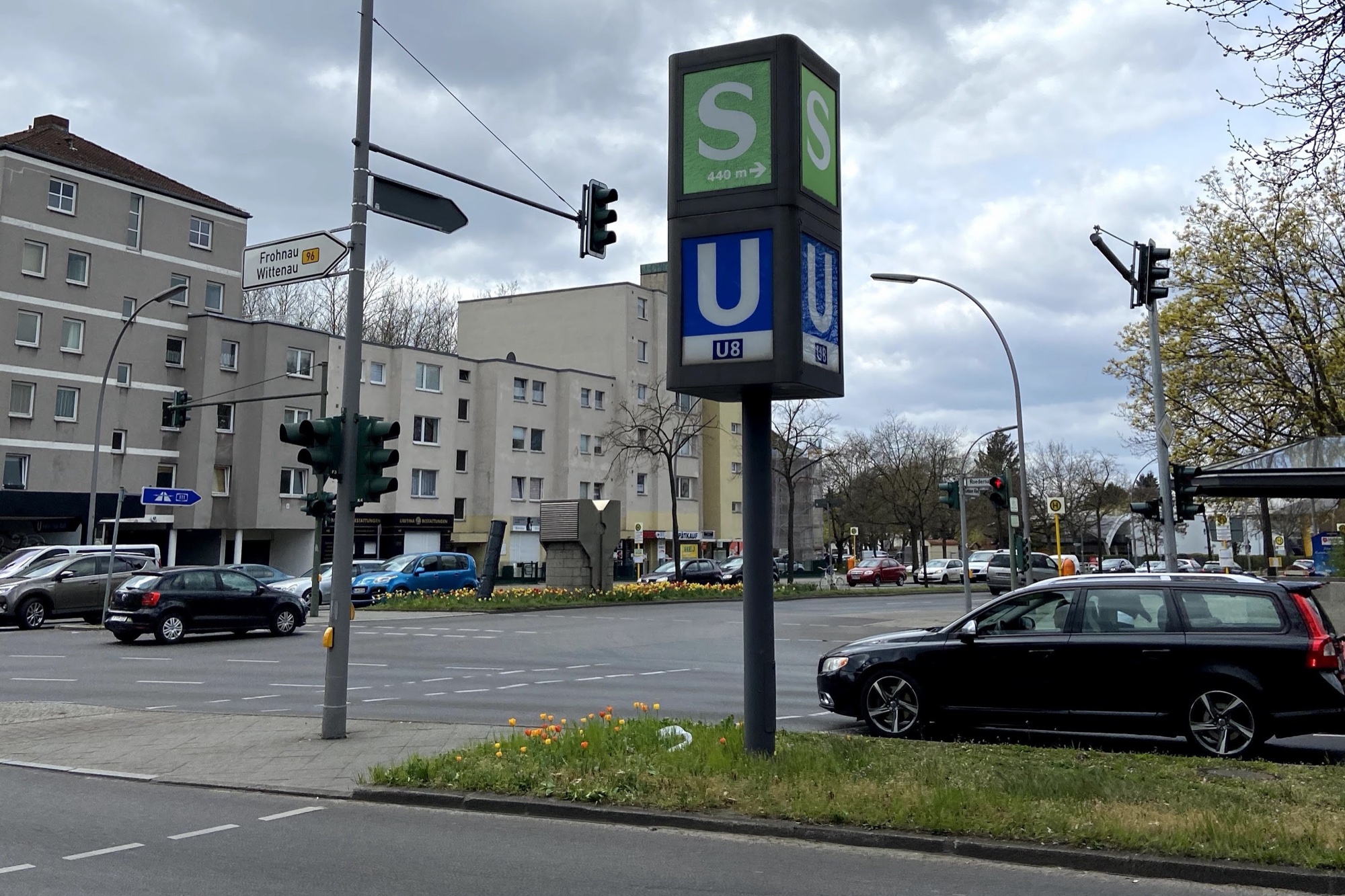 Mehrspurige Ampelkreuzung mit Mittelinsel, auf der eine Wegweiser-Stele für S-Bahn und U-Bahn-Linie U8 steht, dazu Ampeln, Straßenlaternen und parkende Autos, im Hintergrund mehrgeschossige Wohnhäuser
