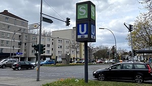 Mehrspurige Ampelkreuzung mit Mittelinsel, auf der eine Wegweiser-Stele für S-Bahn und U-Bahn-Linie U8 steht, dazu Ampeln, Straßenlaternen und parkende Autos, im Hintergrund mehrgeschossige Wohnhäuser