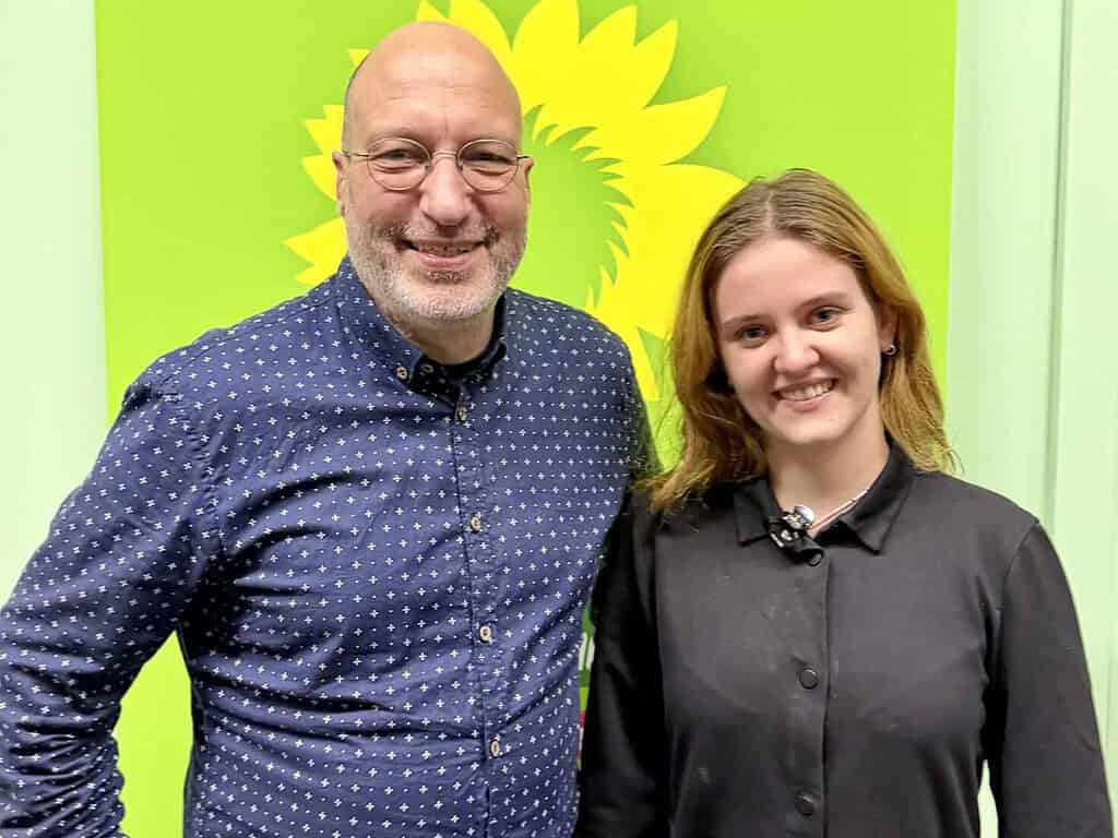 Ein Mann mit Brille und dunkelblauem Hemd und eine junge Frau in schwarzem Kleid stehen lächelnd nebeneinander vor einer grünen Wand mit gelber Sonnenblume, dem Symbol der Grünen