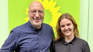 Ein Mann mit Brille und dunkelblauem Hemd und eine junge Frau in schwarzem Kleid stehen lächelnd nebeneinander vor einer grünen Wand mit gelber Sonnenblume, dem Symbol der Grünen