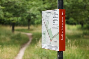 Rot-weiße Orientierungstafel mit der Aufschrift Berliner Mauerweg und einer Karte des Streckenverlaufs an einem Mast, im Hintergrund ein schmaler Pfad durch eine Wiese mit Bäumen.