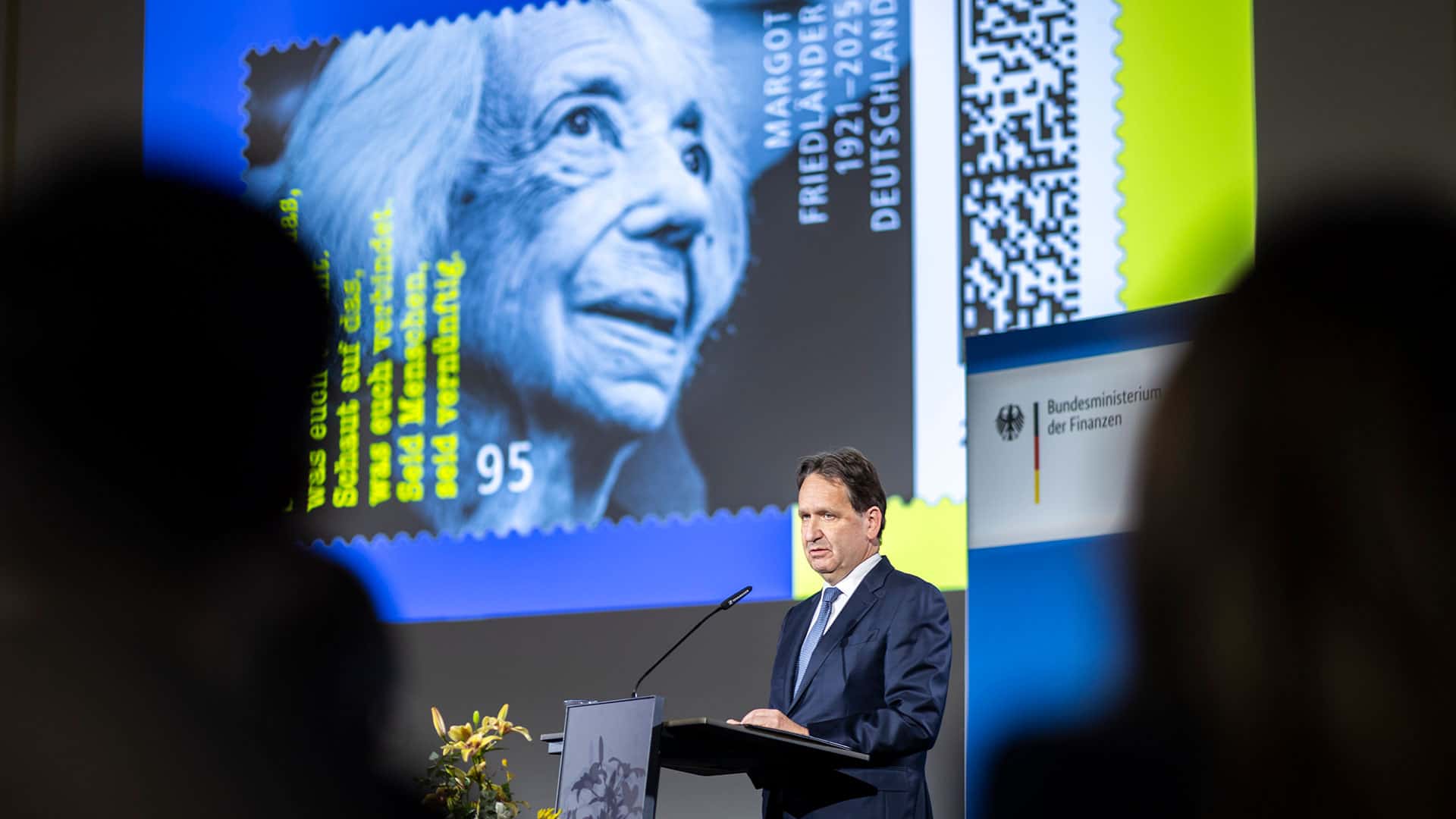 Ein Mann im dunklen Anzug spricht an einem Rednerpult, hinter ihm eine große Projektion einer Briefmarke mit dem Porträt von Margot Friedländer.