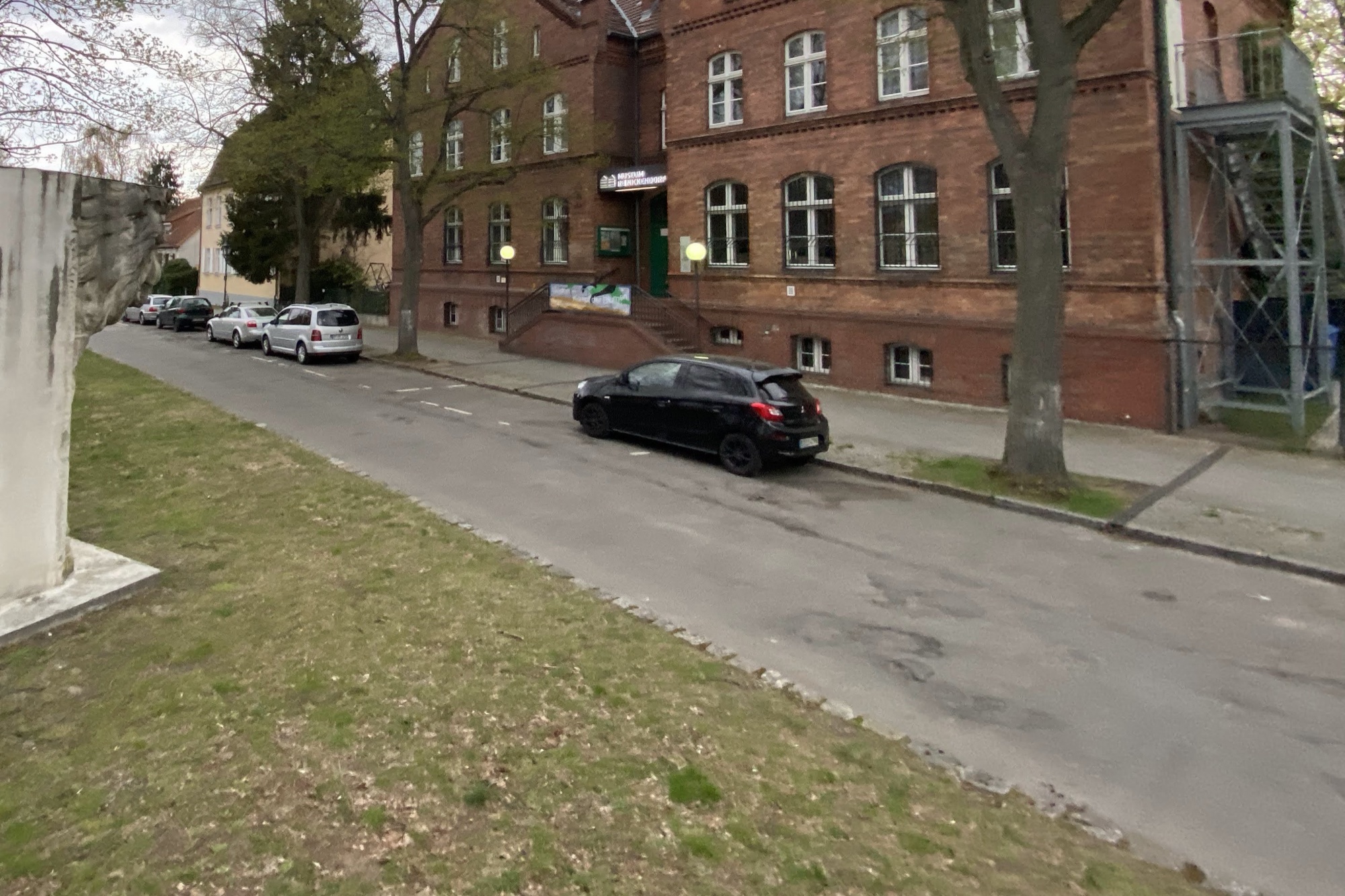 Straße Alt-Hermsdorf mit parkenden Autos vor dem roten Backsteingebäude des Museums Reinickendorf, links eine Steinskulptur auf einer Rasenfläche, kahle Bäume unter bewölktem Himmel