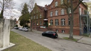 Straße Alt-Hermsdorf mit parkenden Autos vor dem roten Backsteingebäude des Museums Reinickendorf, links eine Steinskulptur auf einer Rasenfläche, kahle Bäume unter bewölktem Himmel