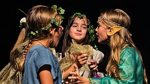 Vier Kinder in Märchenkostümen mit Blätterkränzen, Glitzerstoffen und Elfenohren stehen eng beieinander vor dunklem Hintergrund auf einer Bühne, unten rechts das Logo der Jugendkunstschule ATRIUM.