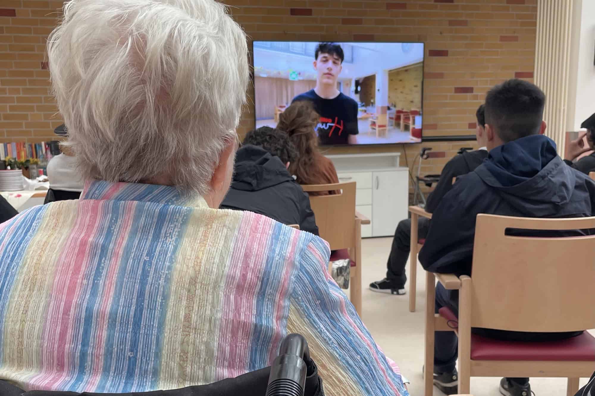 Eine alte Frau und Jugendliche sitzen vor einem Flachbildfernseher auf dem ein Jugendlicher zu sehen ist.