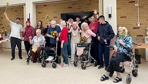 Eine Gruppe aus alten und jungen Menschen winkt in die Kamera, einige halten Blumen in der Hand.