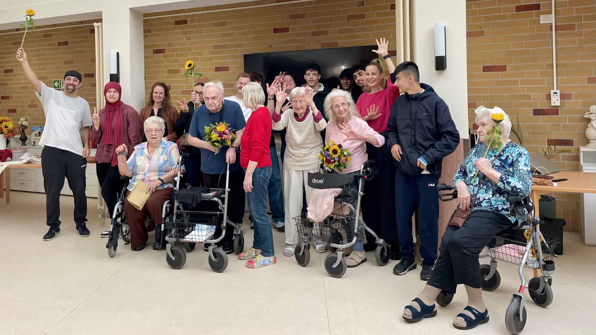 Eine Gruppe aus alten und jungen Menschen winkt in die Kamera, einige halten Blumen in der Hand.