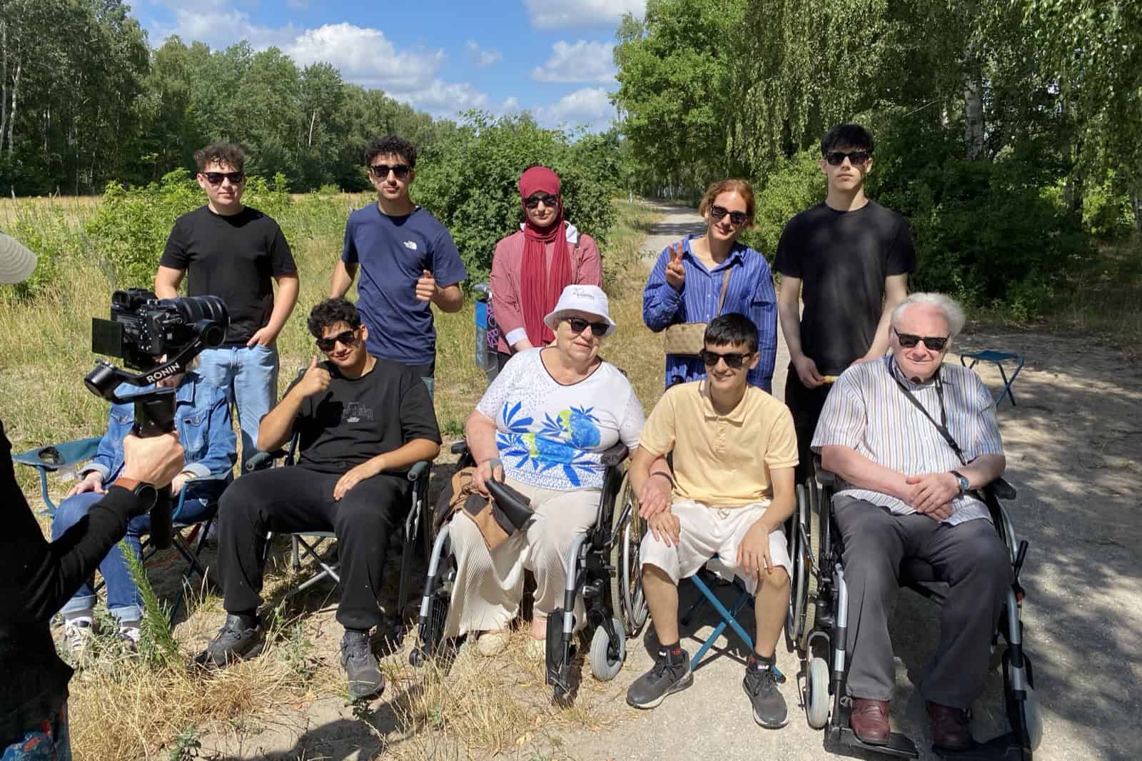 Neun Jugendliche und ältere Menschen sitzen und stehen auf einem Weg im Grünen, links filmt jemand mit einer Kamera.