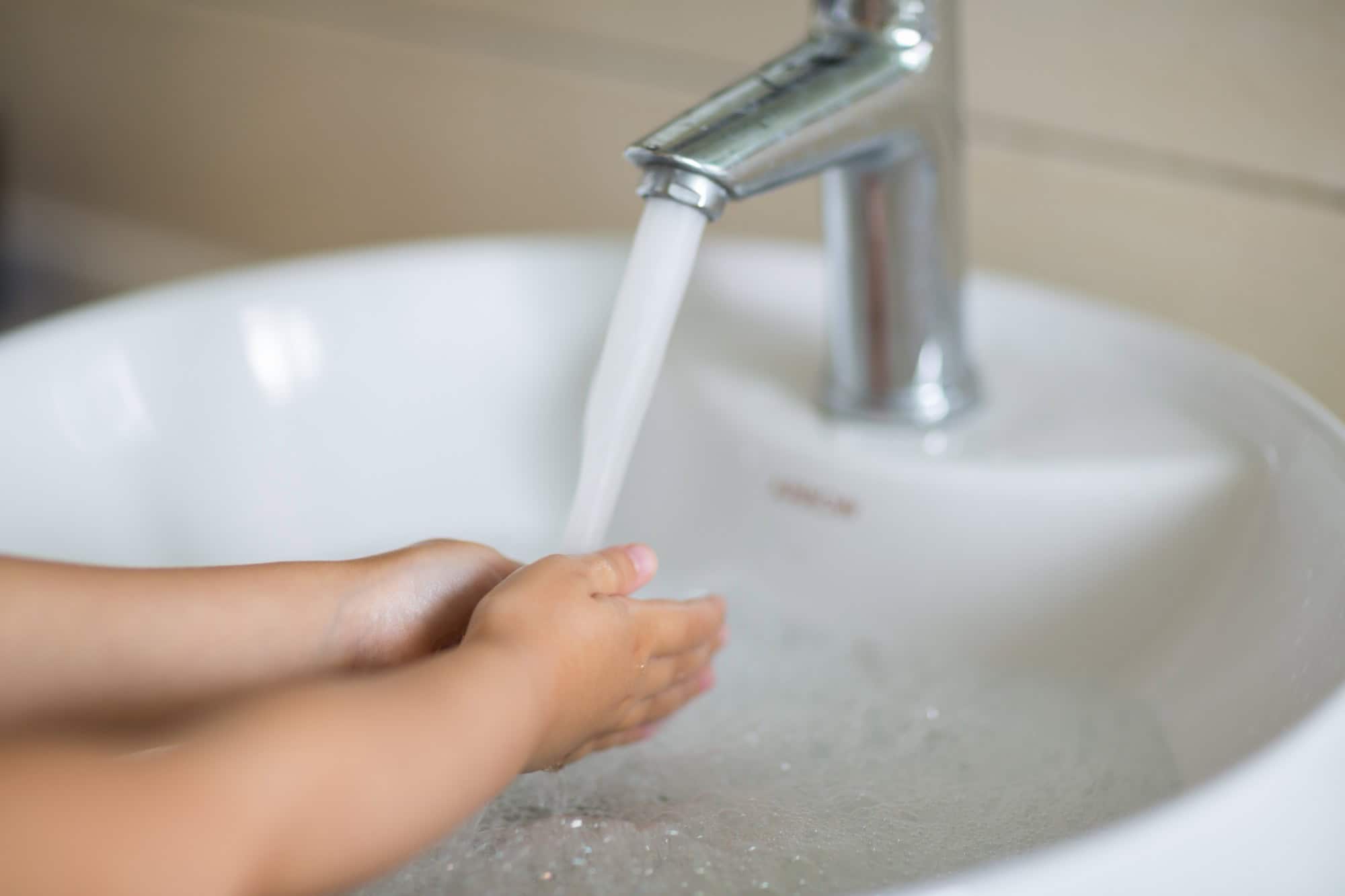Kinderhände unter einem laufenden Wasserhahn über einem weißen Waschbecken.