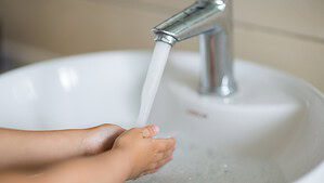 Kinderhände unter einem laufenden Wasserhahn über einem weißen Waschbecken.