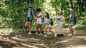 Fünf Kinder mit Rucksäcken wandern durch einen Wald.