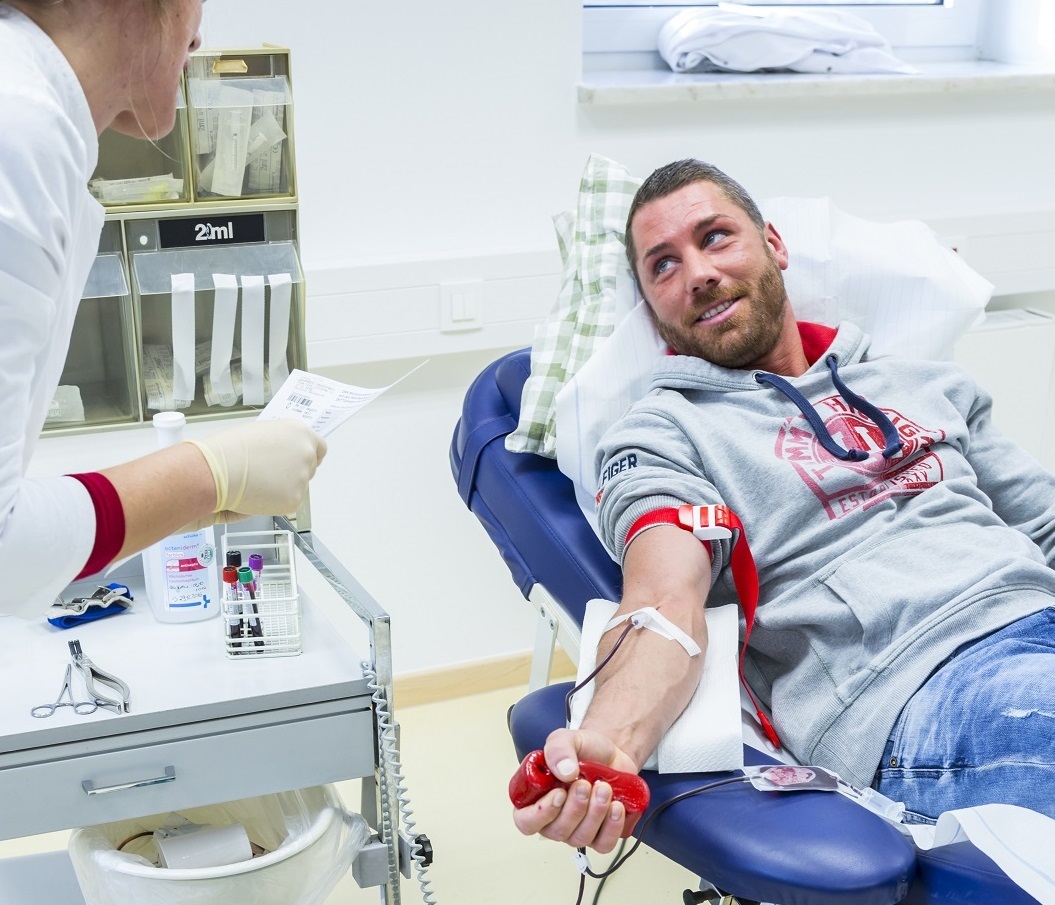 Ein Mann liegt auf einer Liege in einem Behandlungsraum und spendet Blut, während medizinisches Personal neben ihm steht.