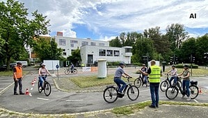 Mehrere Erwachsene mit Fahrradhelmen fahren auf E-Bikes über einen Verkehrsübungsplatz mit Ampeln und Pylonen, in der Mitte gibt ein Trainer in gelber Warnweste Anweisungen, im Hintergrund ein weißes Flachdachgebäude und Bäume