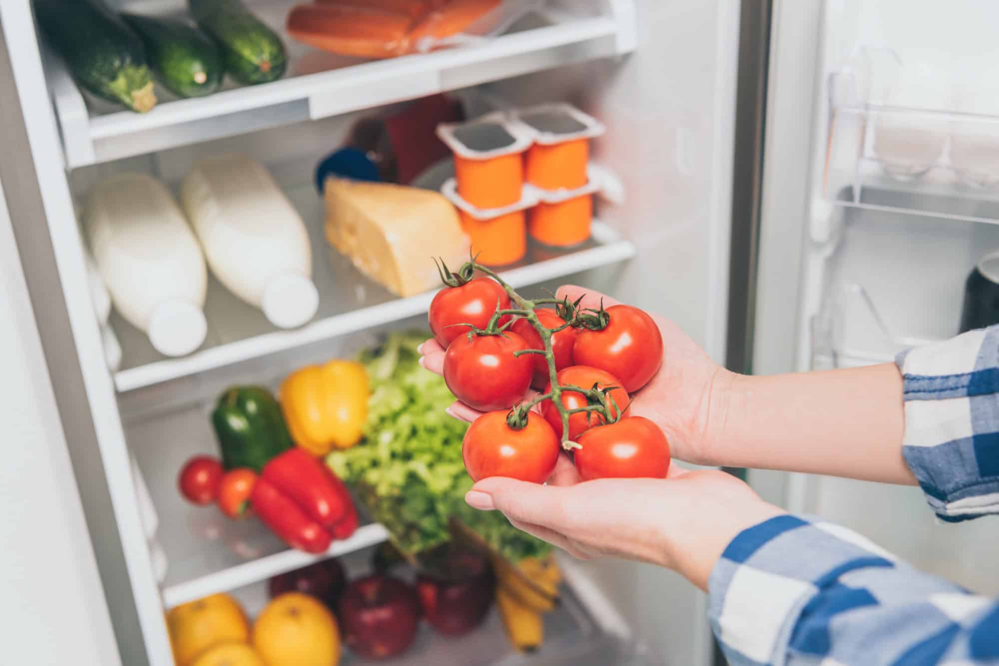 Eine Person legt rote Tomaten in einen gut gefüllten Kühlschrank.