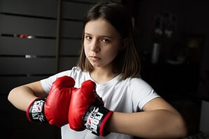Ein jugendliches Mädchen im weißen T Shirt hält in einem Raum rote Boxhandschuhe in die Kamera.